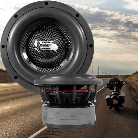 A close-up of two large car subwoofers with a motorcycle parked on the side of an empty road under a cloudy sky in the background.
