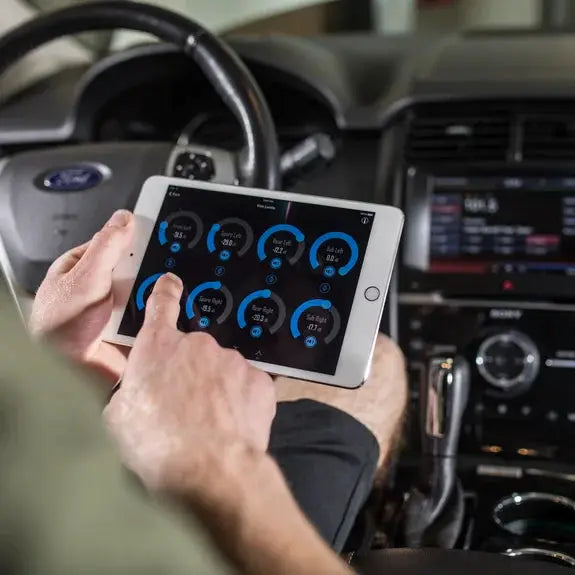 A person sits in a car, holding a tablet displaying diagnostic data from the Rockford Fosgate 8-Channel Interactive Signal Processor with Integrated iDatalink Maestro Module DSR1. The Ford-branded dashboard and steering wheel are visible in the background.