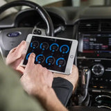 A person sits in a car, holding a tablet displaying diagnostic data from the Rockford Fosgate 8-Channel Interactive Signal Processor with Integrated iDatalink Maestro Module DSR1. The Ford-branded dashboard and steering wheel are visible in the background.